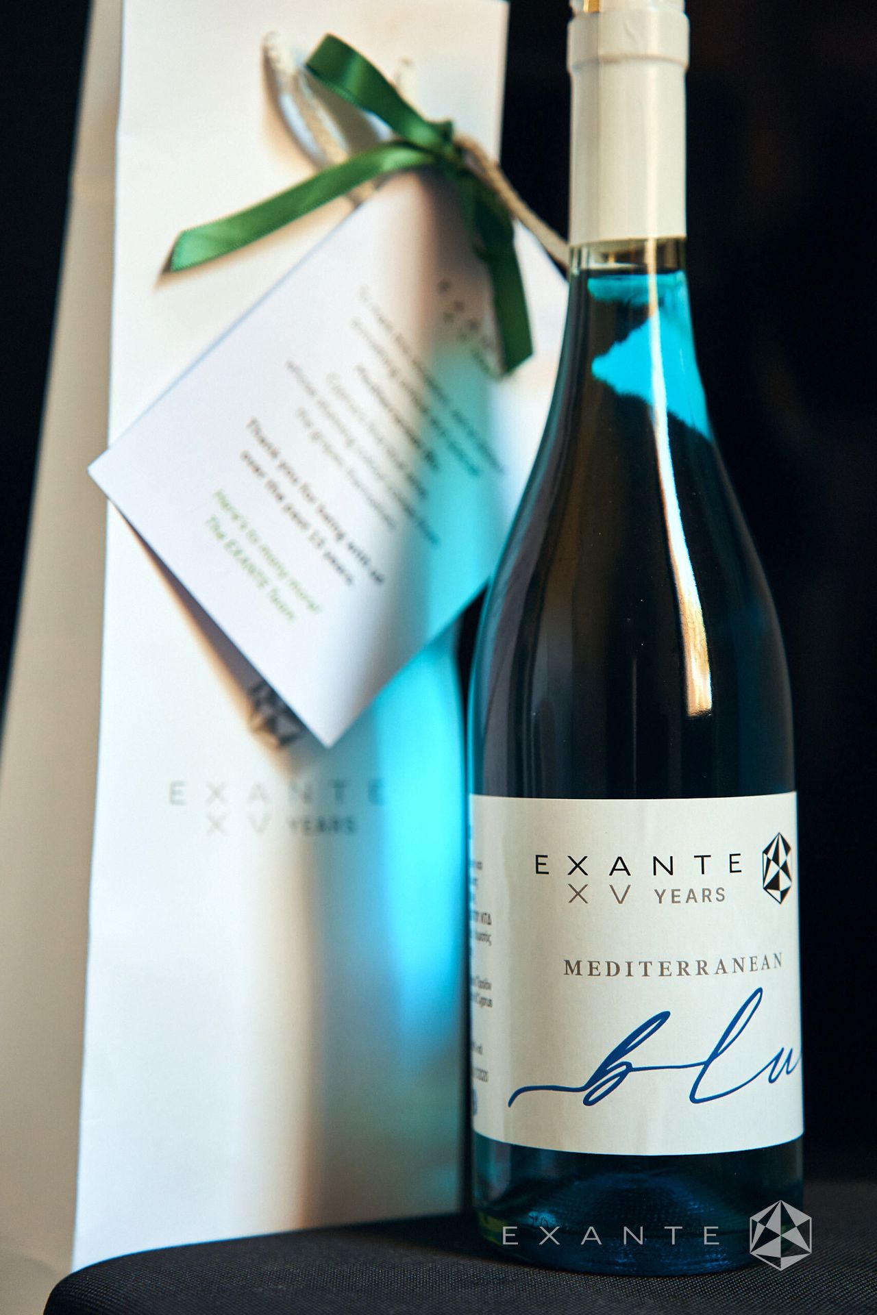 A bottle of Exante XV Years Mediterranean Blue wine stands next to a gift bag with an attached thank-you note.
