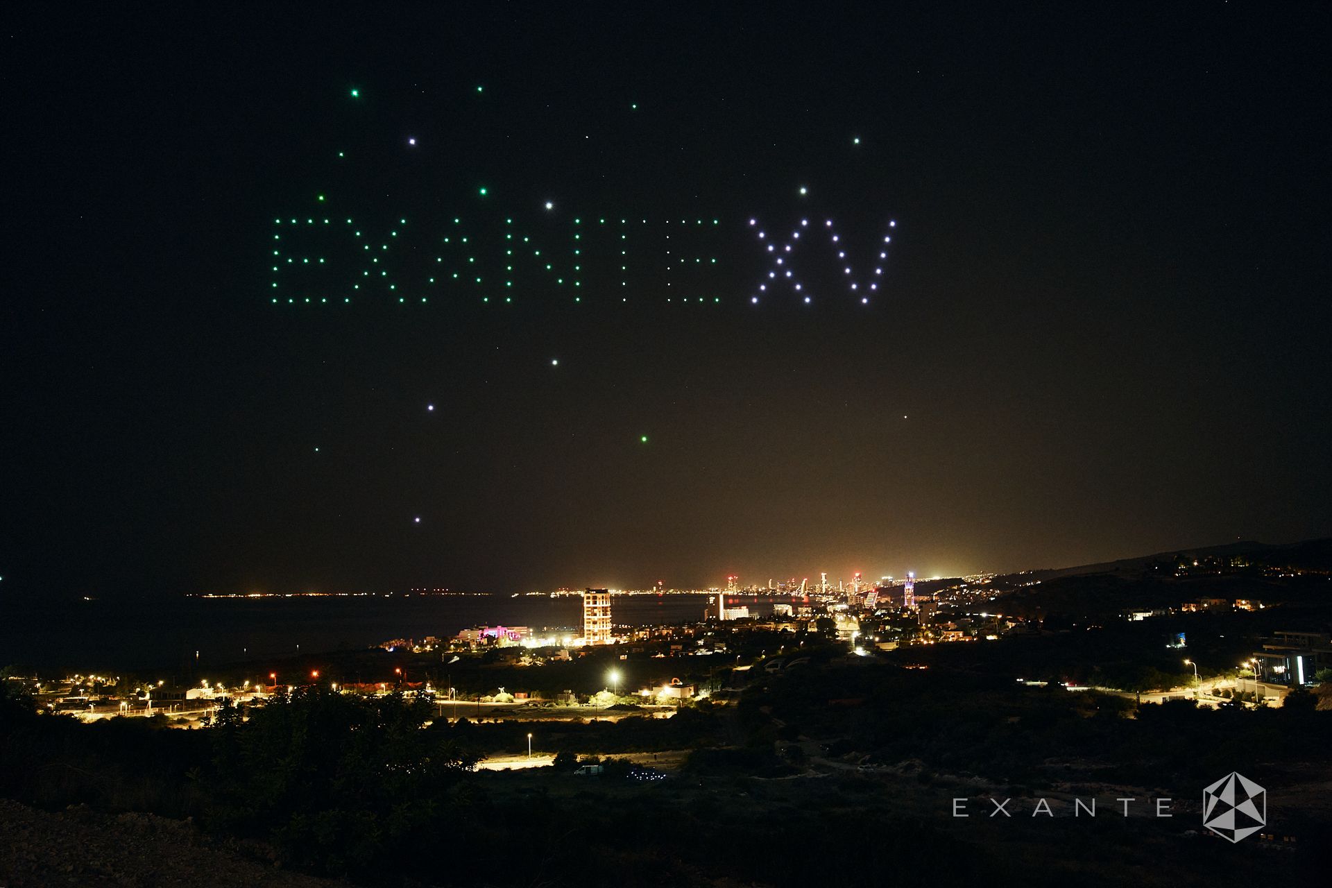 A drone light show displaying the text EXANTE XV over a brightly lit coastal city at night.