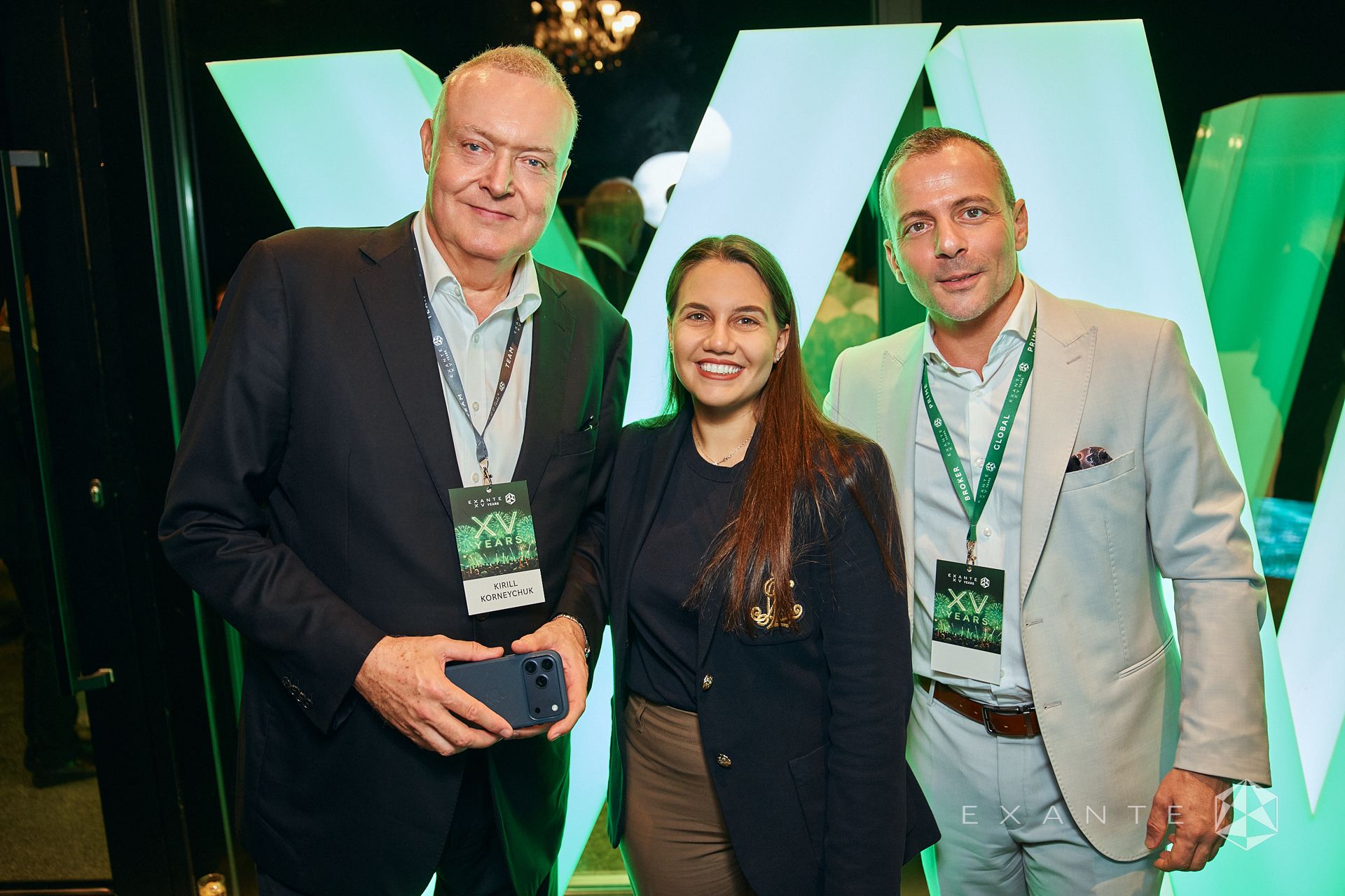 Three people wearing Exante XV Years conference badges pose in front of a large, glowing green W-shaped display.