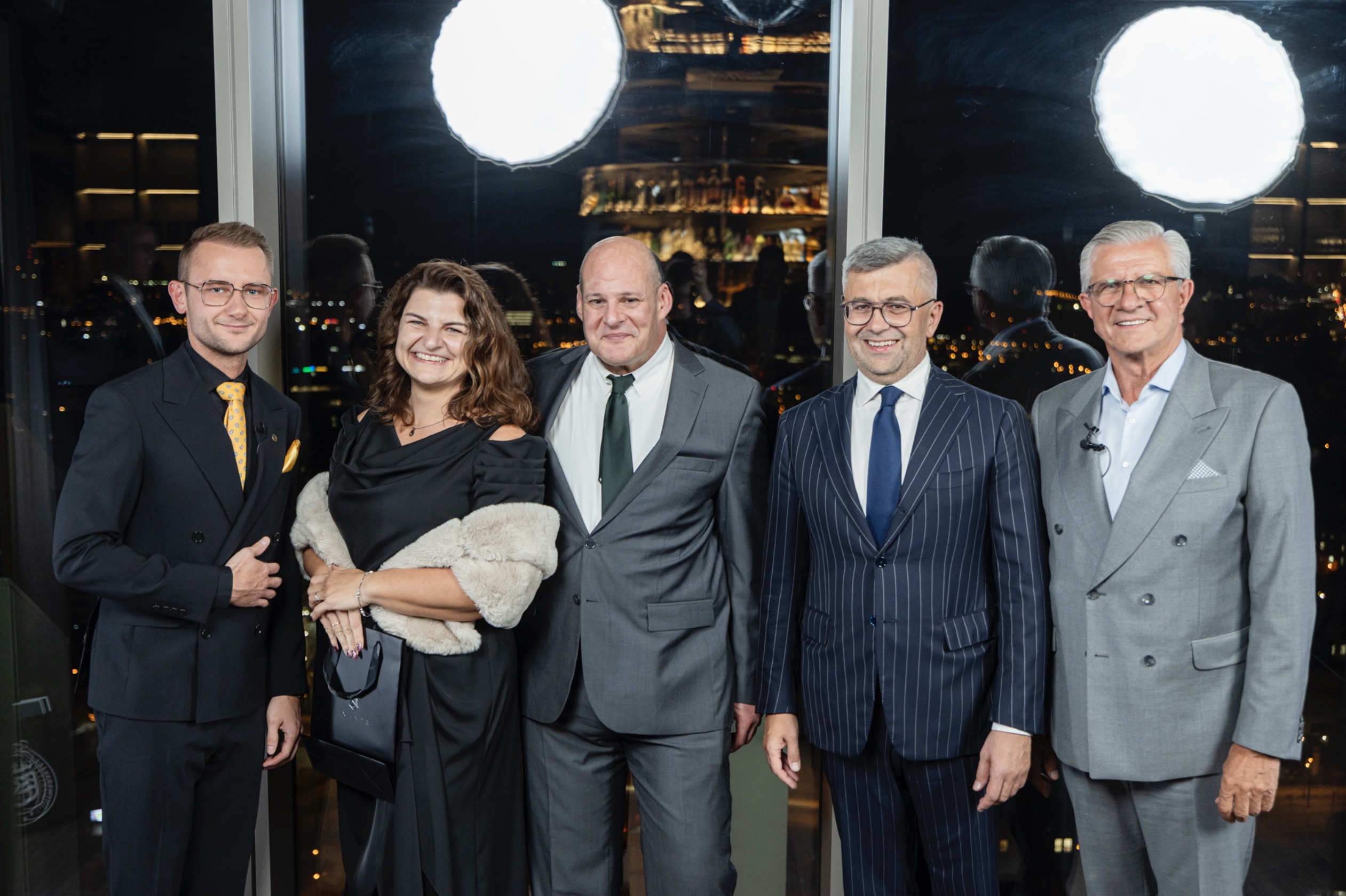 Five people dressed in formal business attire pose together against a night cityscape window background.